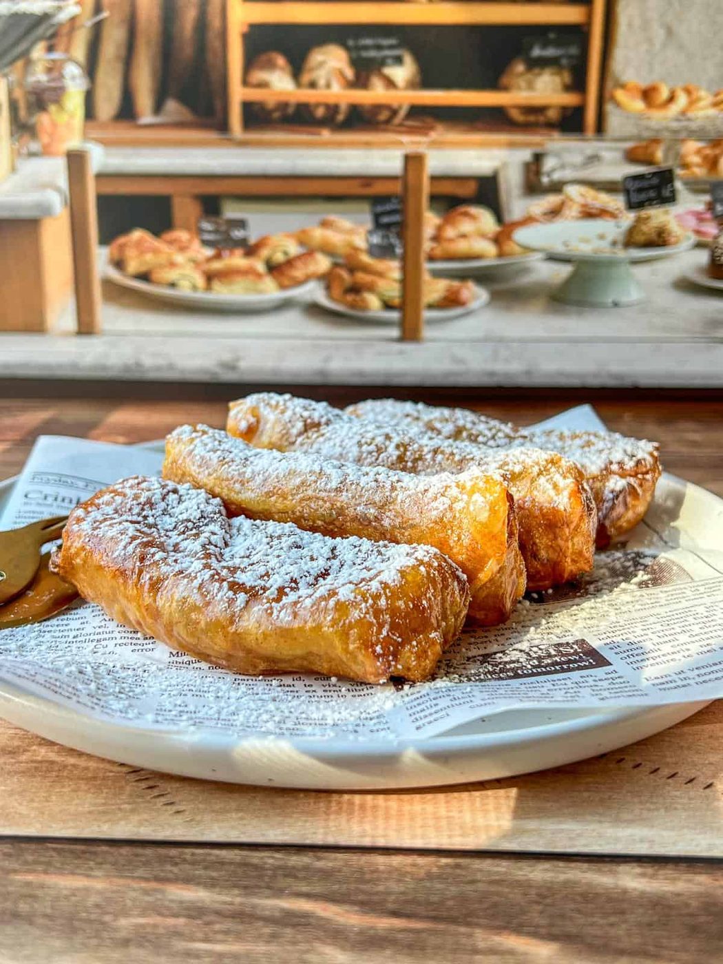 A plate of golden, flaky healthy copycat McDonald's apple pies dusted with powdered sugar sits on a newspaper-lined dish, with more baked goods and pastries visible in the blurred background of a bakery display.