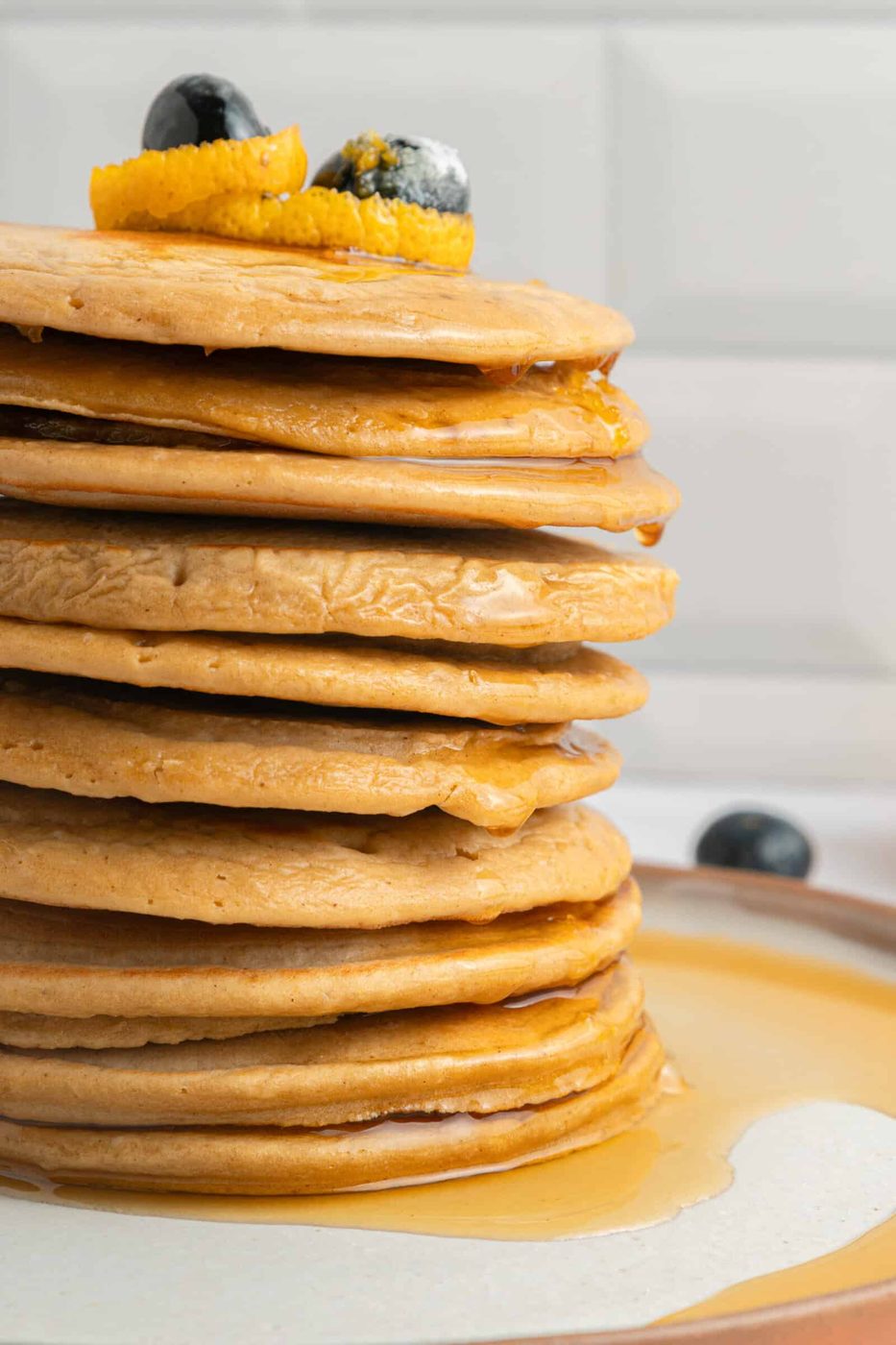 A tall stack of pancakes topped with orange peel and blueberries, with syrup dripping down the sides onto a plate. The background is a white tiled wall.
