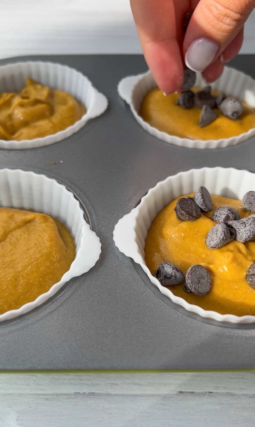A hand sprinkles chocolate chips over batter in silicone muffin cups placed on a baking tray. The batter is a golden color, filling four cups, ready for baking.