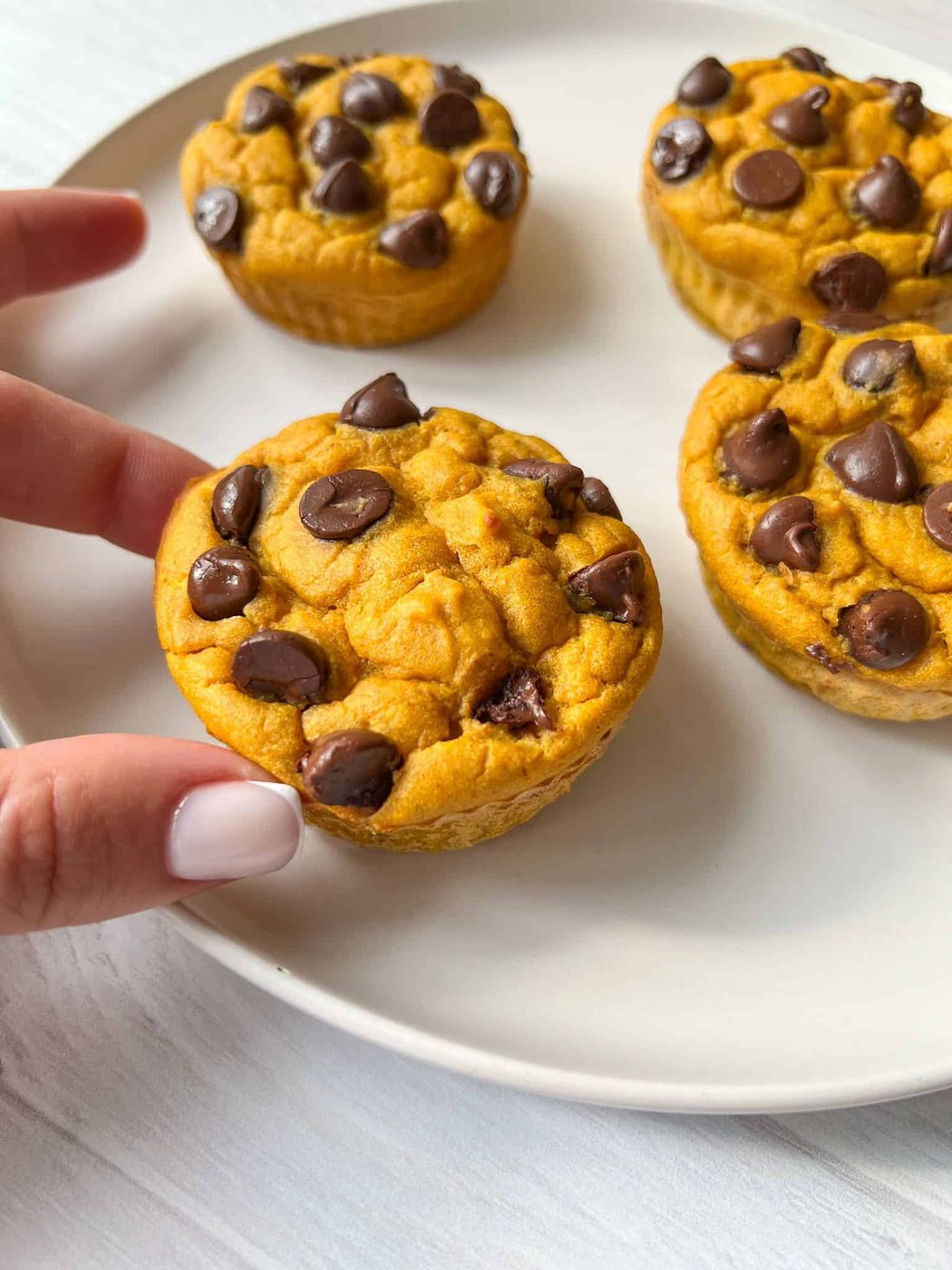 Four chocolate chip muffins are on a white plate. A hand with white nail polish is reaching for one of the muffins. The muffins have a golden-brown color and chocolate chips scattered on top. The background is a light surface.