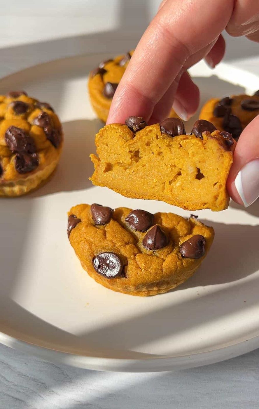 A person holds a half-eaten, round pumpkin muffin with chocolate chips, showing a soft texture. Several whole muffins are on a white plate in the background, illuminated by natural light.