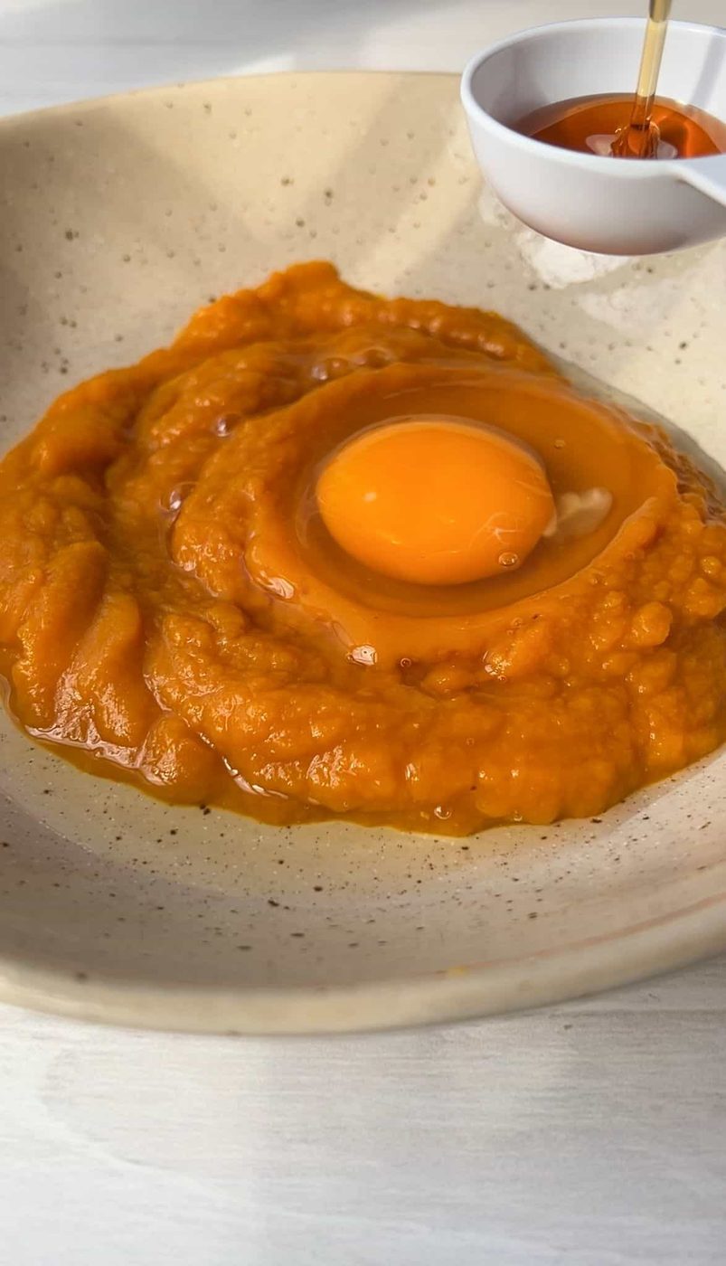 A bowl containing pumpkin puree with a raw egg on top. Honey is being drizzled from a small white bowl above the mixture. The bowl is placed on a light-colored surface.