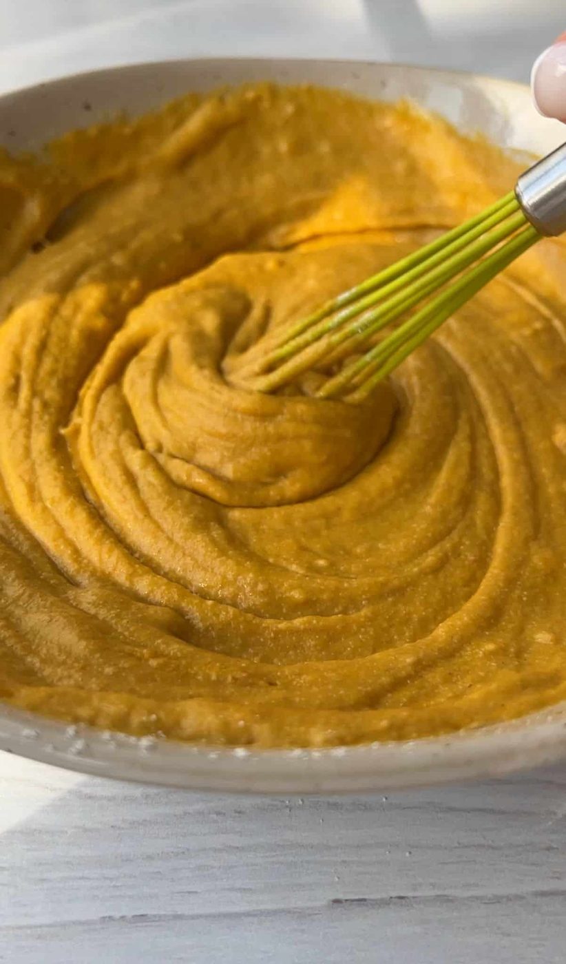A close-up of a creamy, orange-hued mixture being whisked in a beige bowl. The texture appears smooth and thick. The bowl is placed on a light wooden surface.