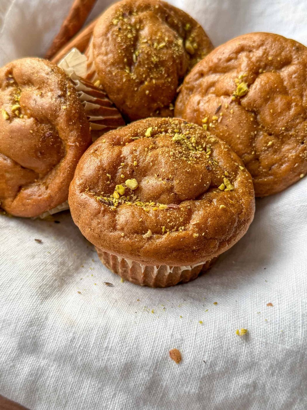 Four golden brown muffins sprinkled with crushed pistachios rest on a white cloth, with one muffin slightly separated in front, showcasing their soft and fluffy texture.