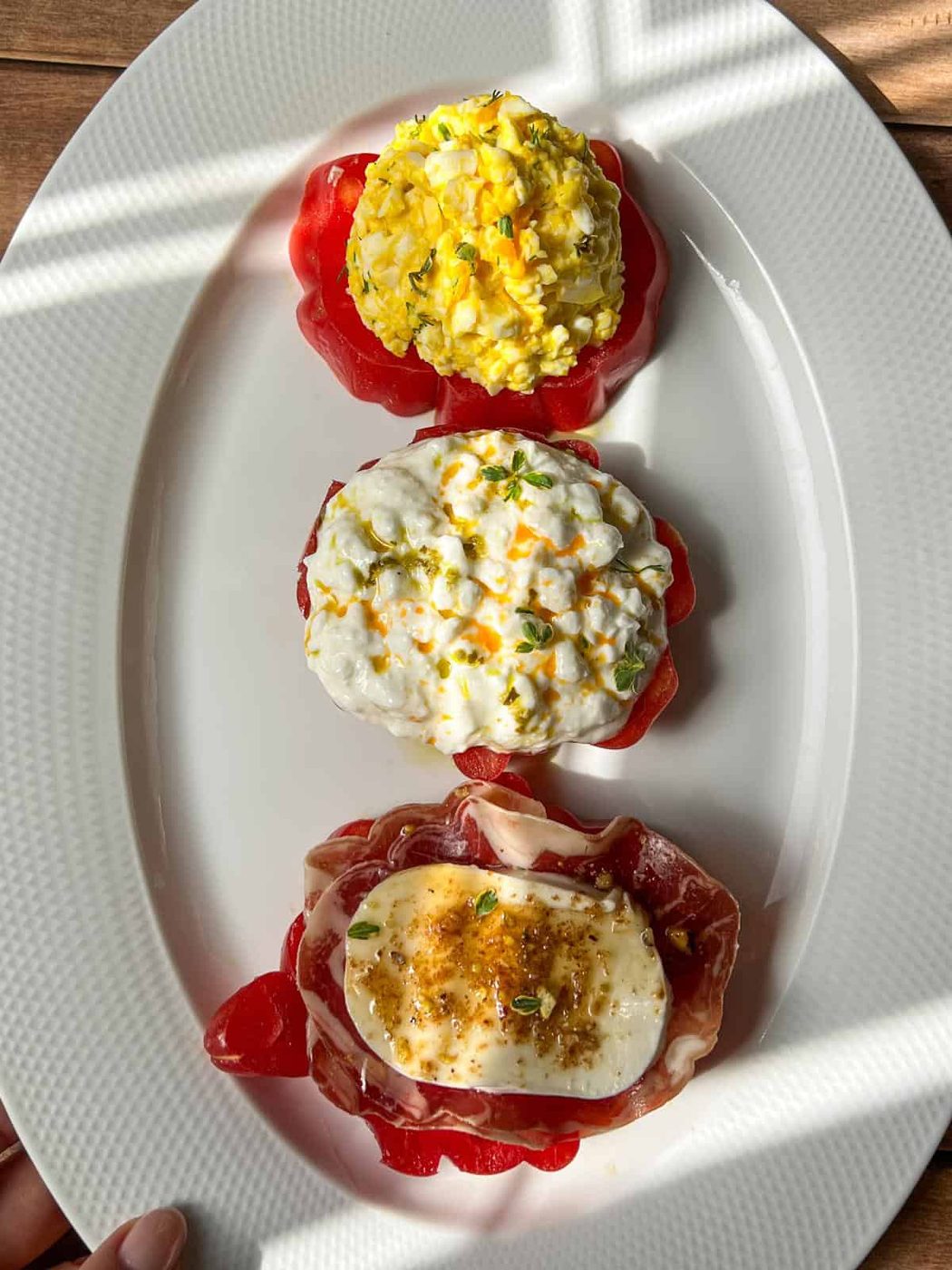Three tomato slices on a white oval plate, each topped with a different mixture: egg salad, cottage cheese with zest, and a slice of mozzarella with cured meat and herbs. Sunlight casts patterned shadows across the dish.