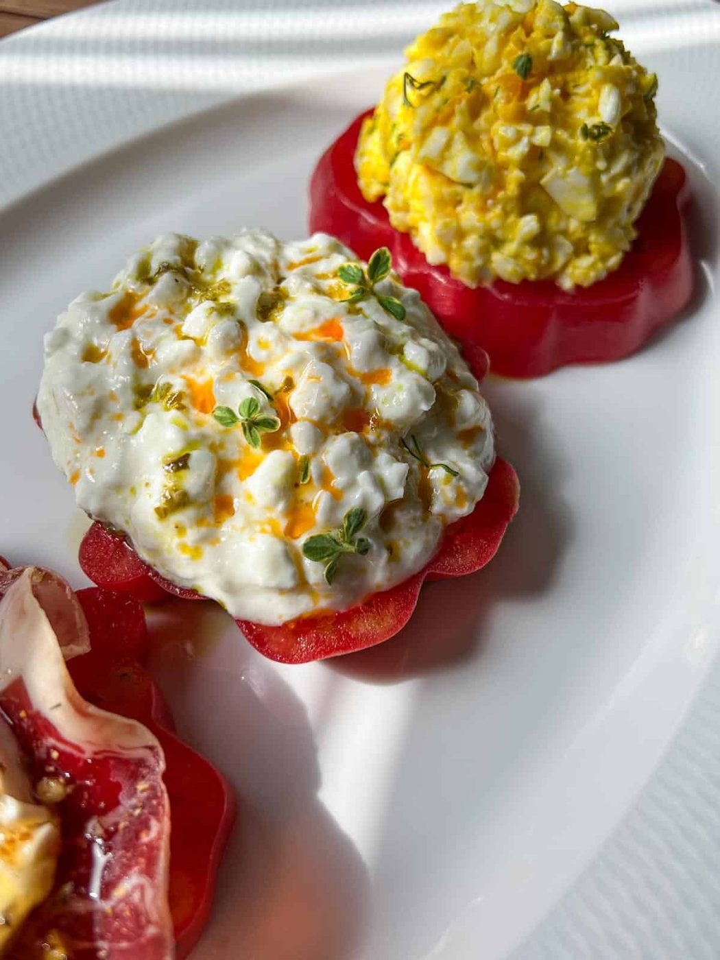 Two tomato slices topped with cottage cheese and egg salad, garnished with fresh herbs, arranged on a white plate. Sunlight highlights the textures and colors of the fresh ingredients.