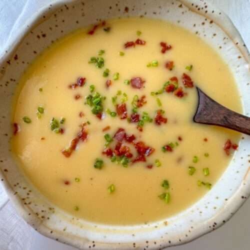 A bowl of creamy yellow soup, reminiscent of Creamy Vegetable Soup, garnished with chopped green herbs and crispy bacon bits, with a wooden spoon resting inside the bowl.