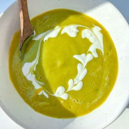 A bowl of Healthy Cream of Broccoli Soup with a swirl of white cream forming a heart-shaped pattern, served with a wooden spoon on the side. Bright natural light casts soft shadows across the bowl.