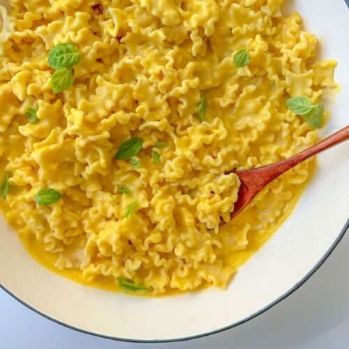 A white bowl filled with ruffled Creamy Butternut Squash Pasta in a silky yellow sauce, garnished with fresh basil leaves, and a wooden spoon resting in the dish.