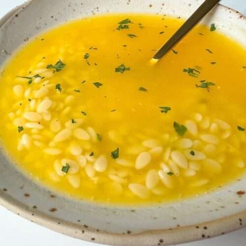 A bowl of yellow Easy Pastina Soup with orzo pasta and sprinkled herbs on top, served in a ceramic bowl with a spoon.