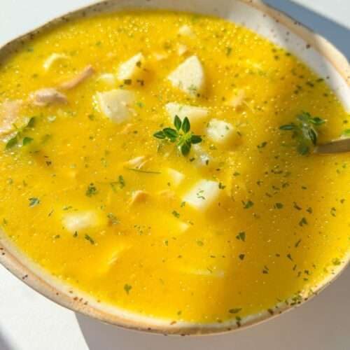 A bowl of Hidden Veggie Chicken Soup with chunks of potato, garnished with herbs, sits on a white surface in bright sunlight.