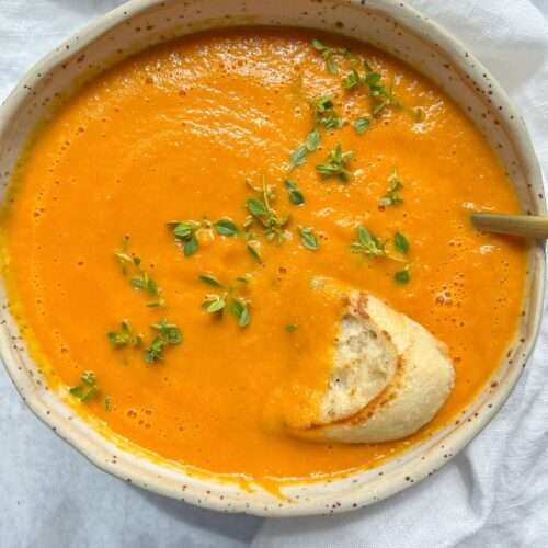 A bowl of creamy tomato soup recipe, vibrant orange and garnished with fresh green herbs, sits invitingly beside a piece of crusty bread partially dipped in the soup on a white cloth background.