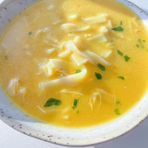 A close-up of a bowl of Hidden Veggie Soup featuring yellow chicken noodle soup with wide noodles, shredded chicken, and sprinkled fresh herbs, served in a light-colored bowl.