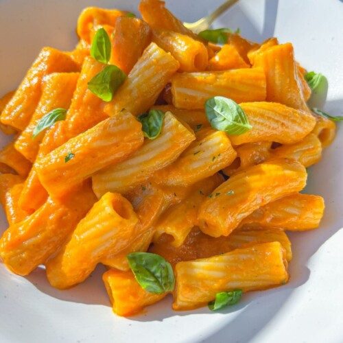 A white plate of rigatoni pasta coated in creamy orange Hidden Veggie Pasta Sauce, garnished with fresh basil leaves, with a gold fork resting on the side.