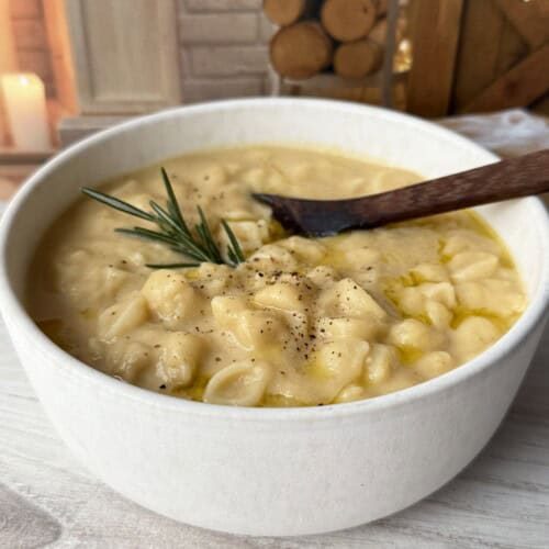 A bowl of creamy pasta with potatoes, garnished with black pepper and a sprig of rosemary, sits on a light wooden surface with a wooden spoon. The cozy background includes stacked firewood and a burning candle.