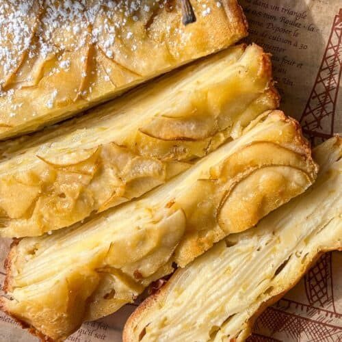 Close-up of several slices of a golden, layered Invisible Pear Cake with thin apple slices visible throughout. The cake is dusted with powdered sugar and placed on a sheet of printed parchment paper.