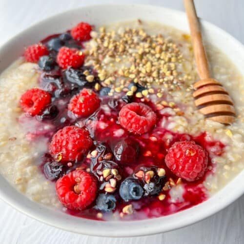 A bowl of oatmeal topped with fresh raspberries, blueberries, a honey drizzle, and a sprinkle of seeds, served with a wooden honey dipper on a light wooden table.