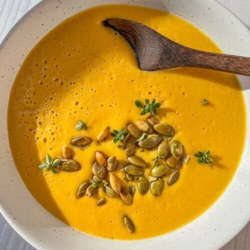 A bowl of creamy orange soup garnished with pumpkin seeds and small green herbs, served with a wooden spoon on a light wooden table.