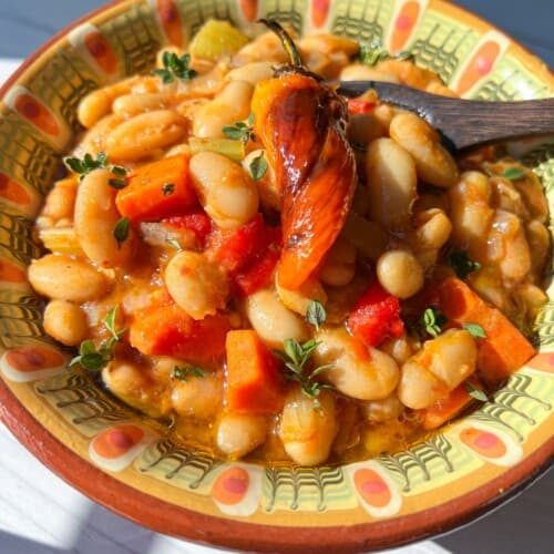 A colorful bowl of Bean Stew with white beans, carrots, red peppers, and herbs, topped with a roasted chili pepper and served in a decorative bowl on a sunlit table.