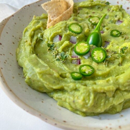 A bowl of creamy homemade guacamole garnished with sliced green chili, fresh herbs, diced red onion, and served with tortilla chips on the side.