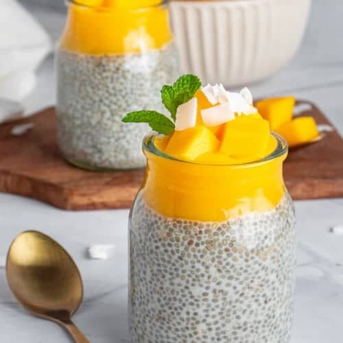 Two glass jars filled with chia pudding topped with mango chunks, coconut flakes, and mint leaves. A gold spoon lies beside them. In the background, a bowl of mangoes sits on a wooden board.