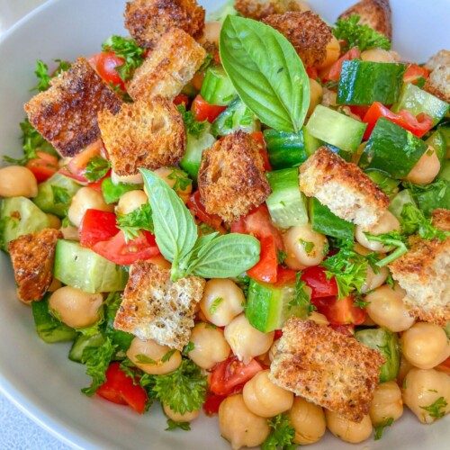 A bowl of Chickpea Crouton Salad with diced cucumbers, tomatoes, fresh parsley, crunchy croutons, and basil leaves, all beautifully arranged in a white bowl.