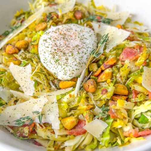 A white bowl filled with shaved Brussels sprouts, pistachios, bacon pieces, Parmesan shavings, herbs, and a poached egg on top—this Julia Roberts Salad is garnished with cracked pepper for a fresh twist.