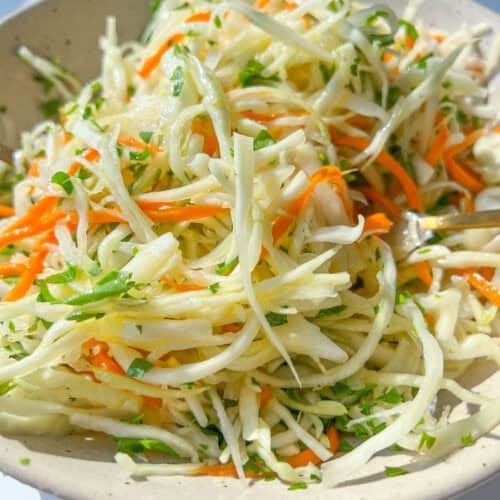 A close-up of a bowl filled with fresh coleslaw made of shredded cabbage, carrots, and parsley in a light dressing. This Healthy Coleslaw Recipe is bright and colorful, served in a beige bowl on a white surface.