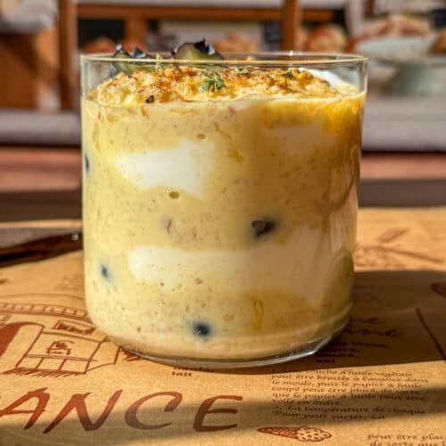 A glass filled with layers of creamy yogurt, yellow pudding, and blueberries, topped with granola and herbs, placed on a decorative placemat with bakery shelves blurred in the background.