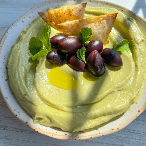 A bowl of creamy Avocado Hummus topped with black olives, fresh mint leaves, a drizzle of olive oil, and two seasoned tortilla chips sits on a light wooden surface in natural sunlight.