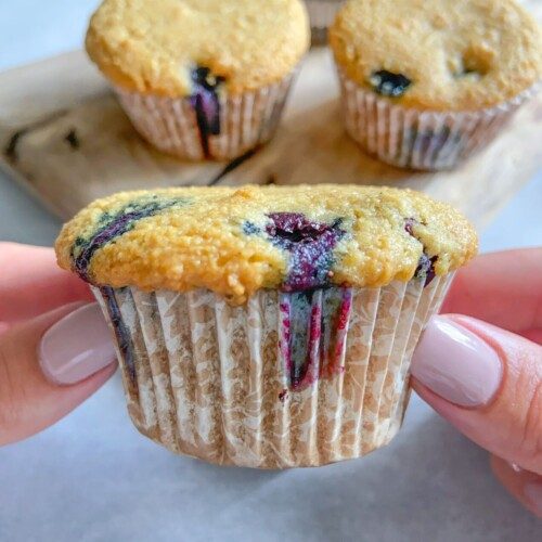 A person with light-painted nails holds an Almond Flour Blueberry Muffin in a paper wrapper, with three more muffins on a wooden board in the background. The muffin has visible blueberries and a golden-brown top.