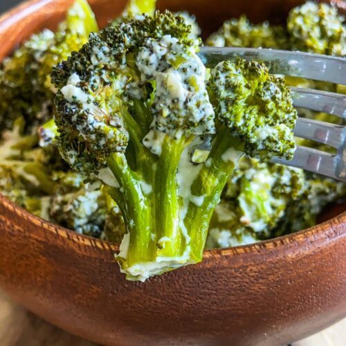 A close-up of Creamy Baked Broccoli florets coated in a luscious white sauce, with a fork lifting one piece above a wooden bowl brimming with more broccoli.