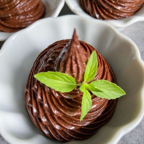 A white bowl filled with swirled chocolate mousse, topped with a small sprig of fresh green mint. Hints of crushed Peanut Butter Cups add texture, while two other similar bowls are partially visible in the background.