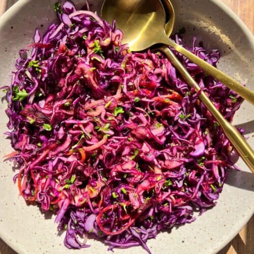 A bowl of colorful red cabbage coleslaw garnished with parsley is served in a light-colored dish. Two gold-colored utensils are placed on the side. The dish is set on a wooden surface, illuminated by natural light.