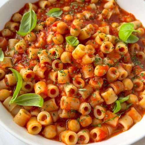 A white bowl filled with ditalini pasta in a rich tomato sauce, inspired by Pasta e Ceci, garnished with fresh basil leaves and sprinkled with herbs.
