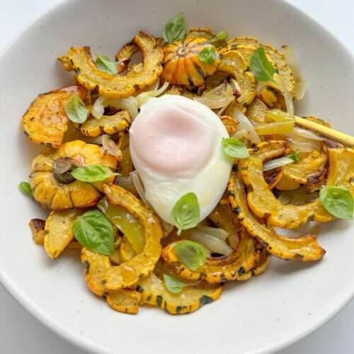 A white plate features a Delicata Squash Salad with roasted delicata squash slices, sautéed onions, fresh basil leaves, and a poached egg in the center, all artfully arranged on a white background.