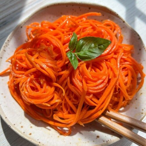 A plate of spiralized carrot noodles inspired by Korean Carrot Salad is garnished with a fresh basil leaf, with chopsticks resting on the side. Sunlight casts gentle shadows across the light-colored wooden table.