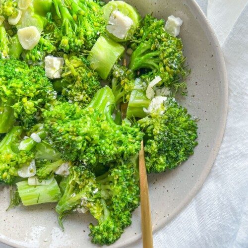 A bowl of fresh sauteed broccoli with feta, mixed with sliced garlic and small cubes of white cheese, served on a light-colored plate with a gold fork resting inside.