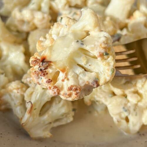Close-up of a bowl of creamy roasted cauliflower on a beige plate. A fork is holding a piece of cauliflower, displaying a golden-brown edge, with sauce and herbs visible. The dish is set on a light-colored surface.