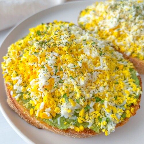 A close-up of toast topped with a layer of avocado and finely grated hard-boiled egg, garnished with herbs, alongside crispy Pasta Chips on a white plate.