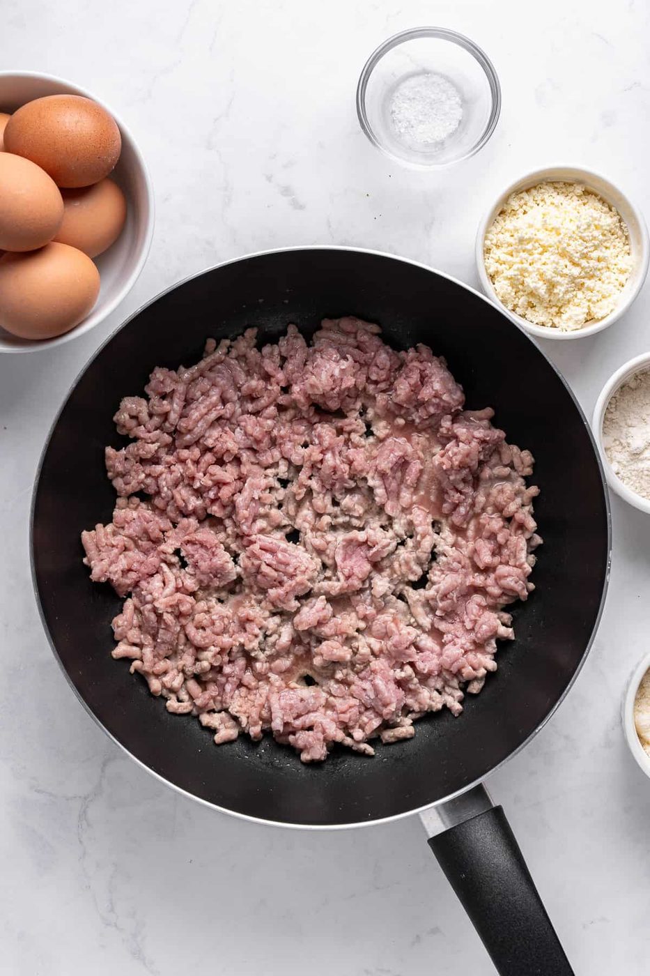 A frying pan with raw ground meat, surrounded by small bowls containing eggs, flour, breadcrumbs, and salt on a white marble surface.