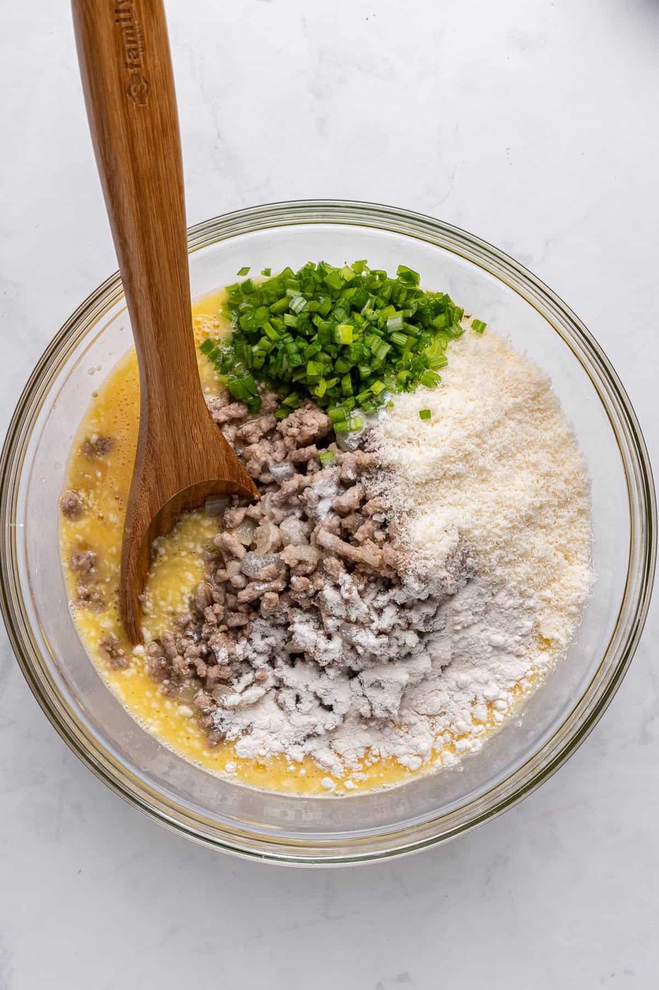 A glass bowl with grated cheese, chopped green onions, cooked sausage, and flour atop a mixture of beaten eggs. A wooden spoon is placed inside the bowl, ready to mix the ingredients. The bowl rests on a white marble surface.