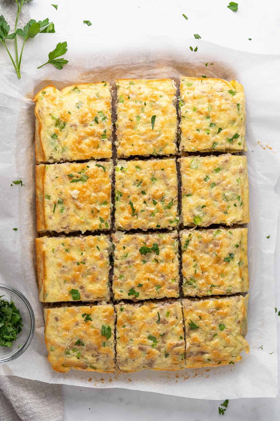 Golden, cheesy baked squares topped with chopped parsley on parchment paper. A small bowl of parsley is on the side, adding a touch of freshness to the presentation.