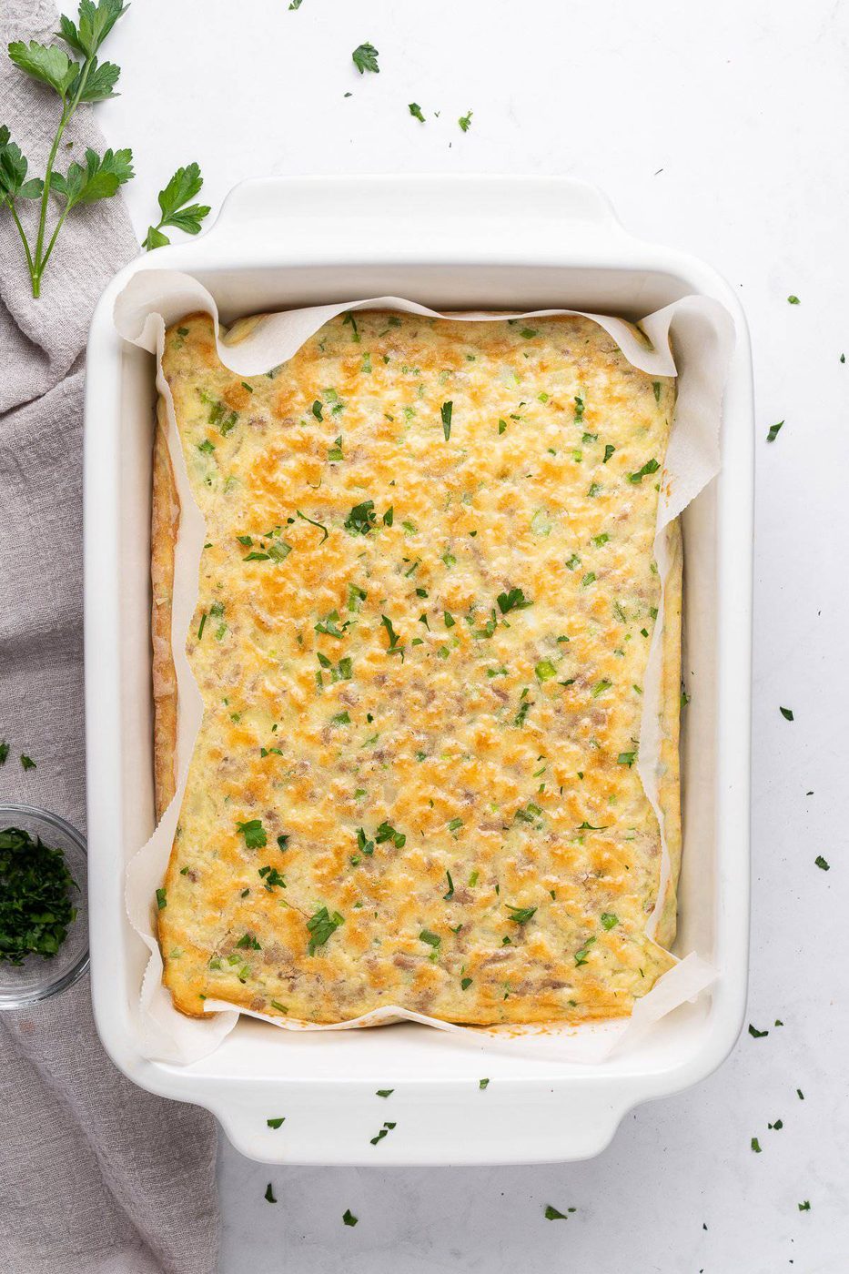 A freshly baked high protein breakfast casserole in a rectangular white dish. The casserole is topped with melted cheese and sprinkled with chopped herbs. A gray cloth and a small bowl of herbs are nearby on the white countertop.