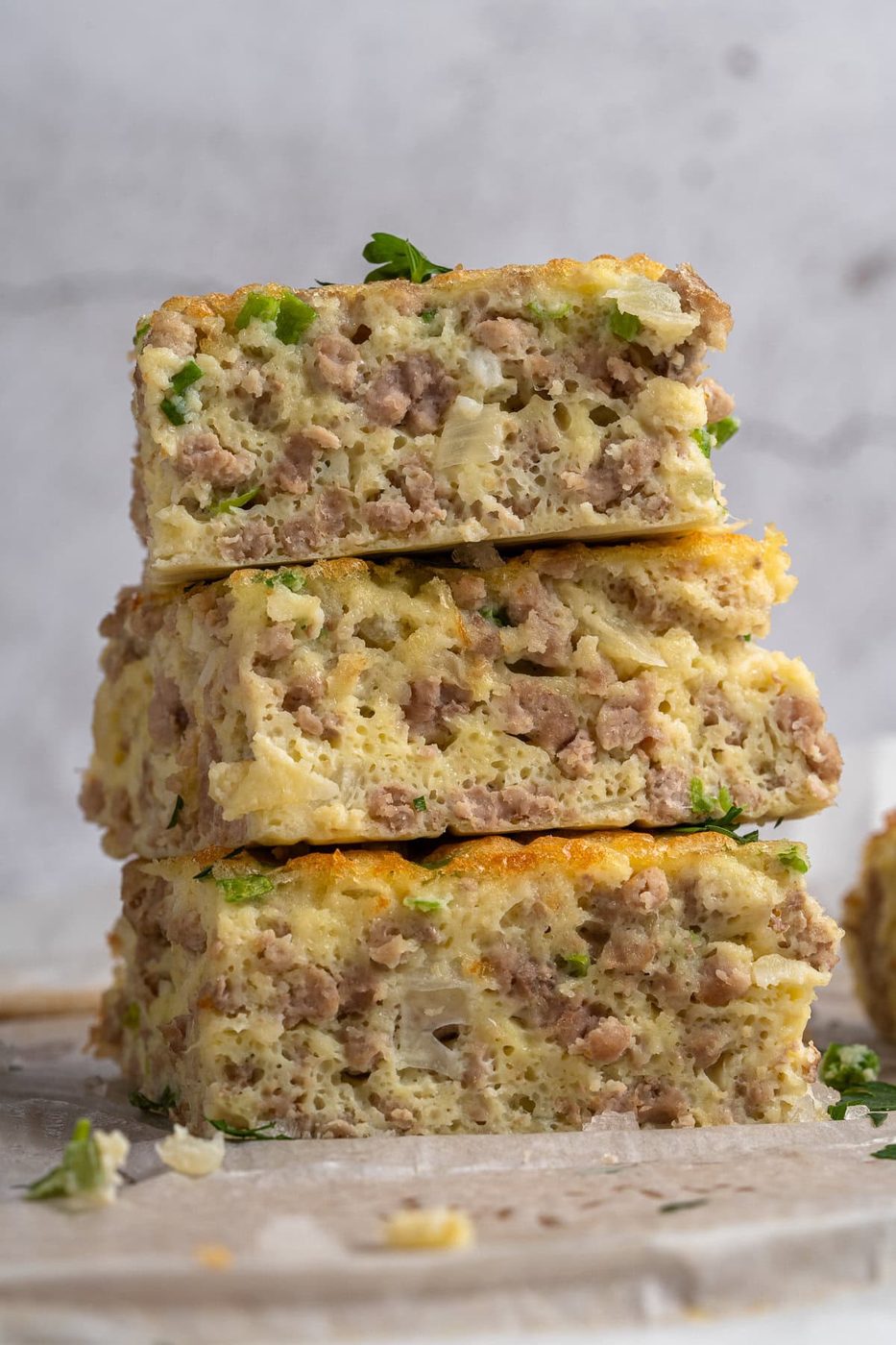 Three slices of savory breakfast casserole stacked vertically, filled with sausage, eggs, and green herbs. The soft, fluffy texture is visible, and there are sprinkles of parsley on top. The background is a light stone surface.