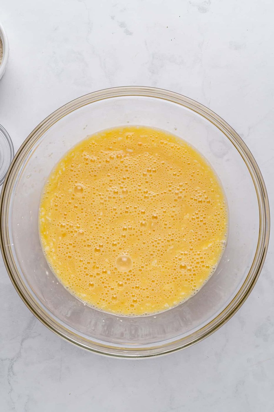A glass bowl filled with beaten eggs, creating a frothy yellow mixture. The bowl rests on a light gray marbled surface.