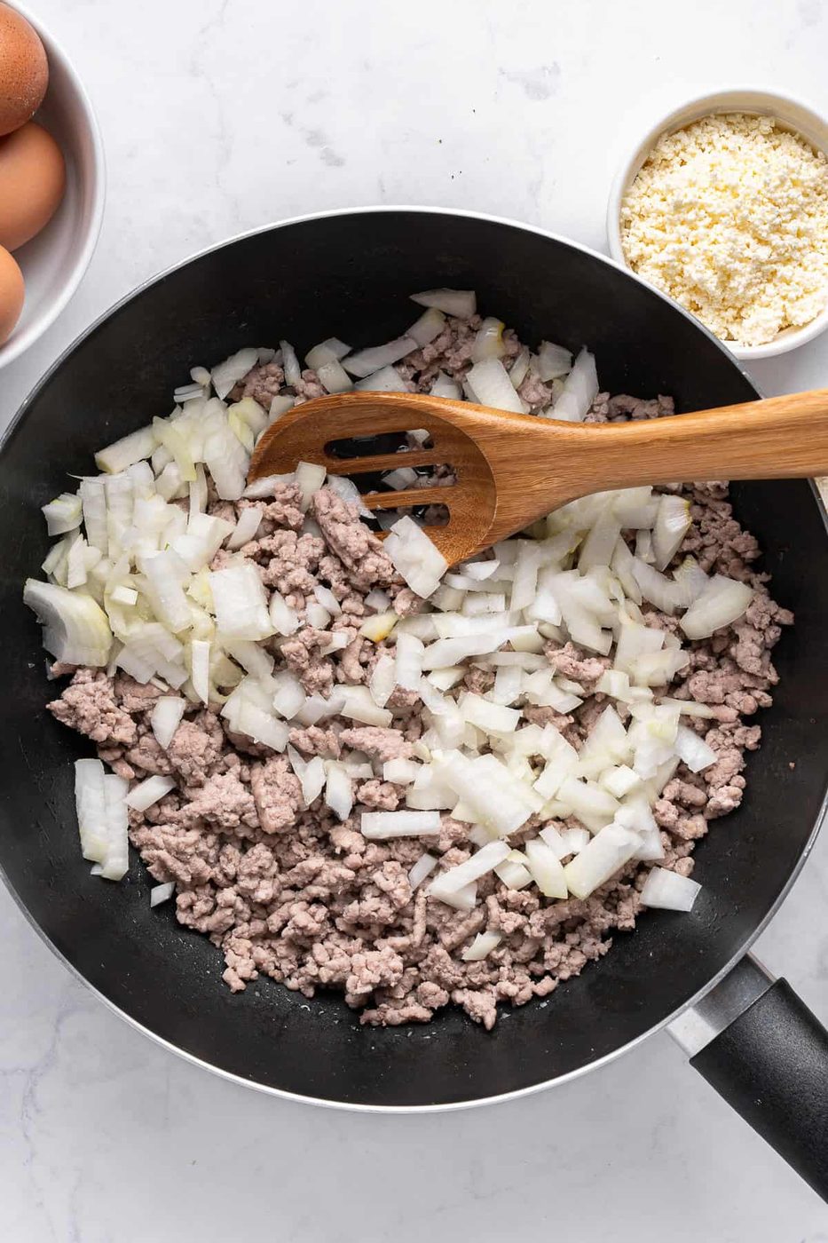 A frying pan on a white surface contains minced meat and chopped onions, being stirred with a wooden spatula. Surrounding the pan are eggs and a bowl of grated cheese.