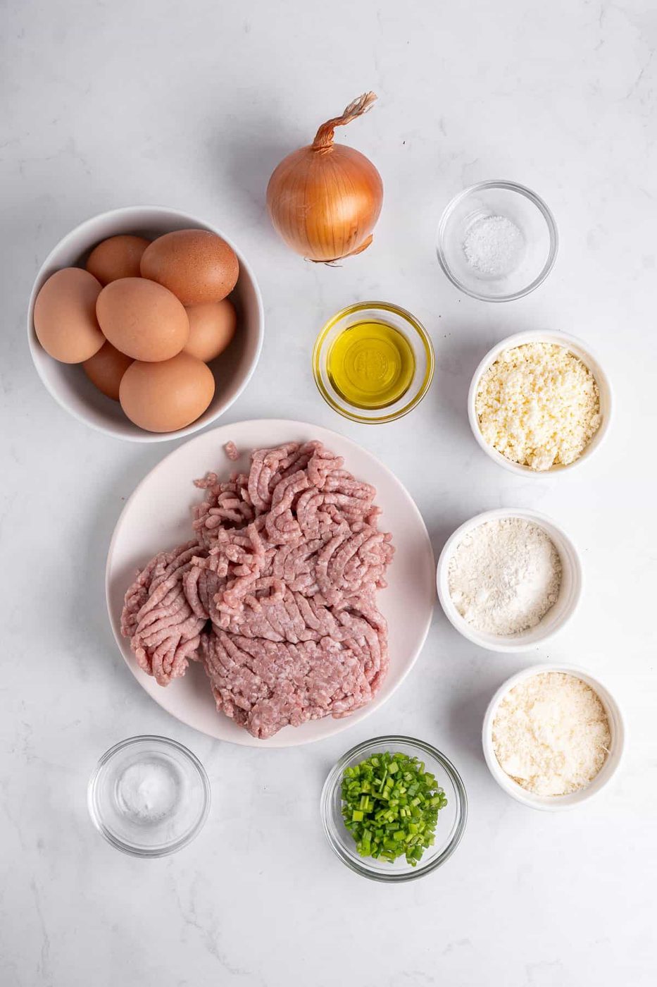Top-down view of raw ingredients: a bowl of eggs, an onion, ground meat on a plate, small bowls of olive oil, flour, breadcrumbs, grated cheese, chopped green onions, and salt. All items are arranged on a white surface.
