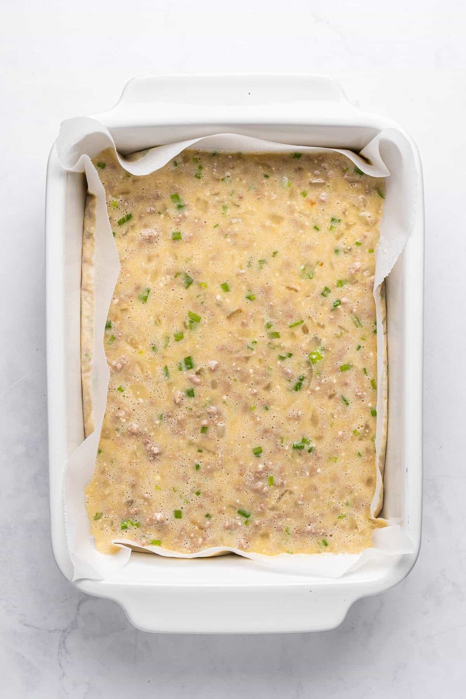 Overhead view of an uncooked egg and vegetable mixture in a parchment-lined white baking dish. The mixture includes visible green herbs and bits of vegetables.