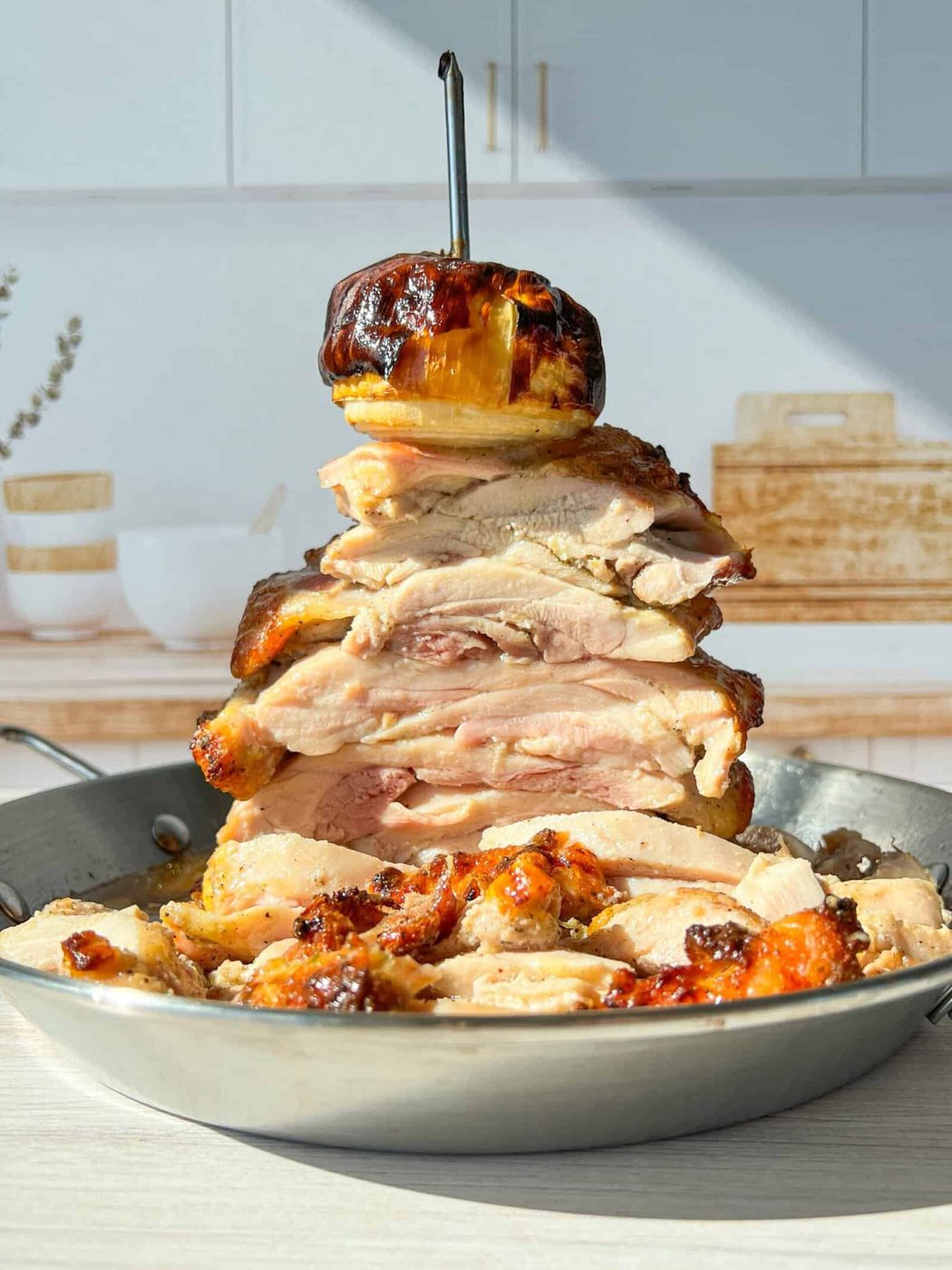 High Protein Chicken Gyro. A towering stack of sliced, roasted chicken pieces is skewered on a vertical rod in a pan. A roasted onion tops the stack. The setting includes a bright kitchen with a wooden cutting board and decorative jars in the background.